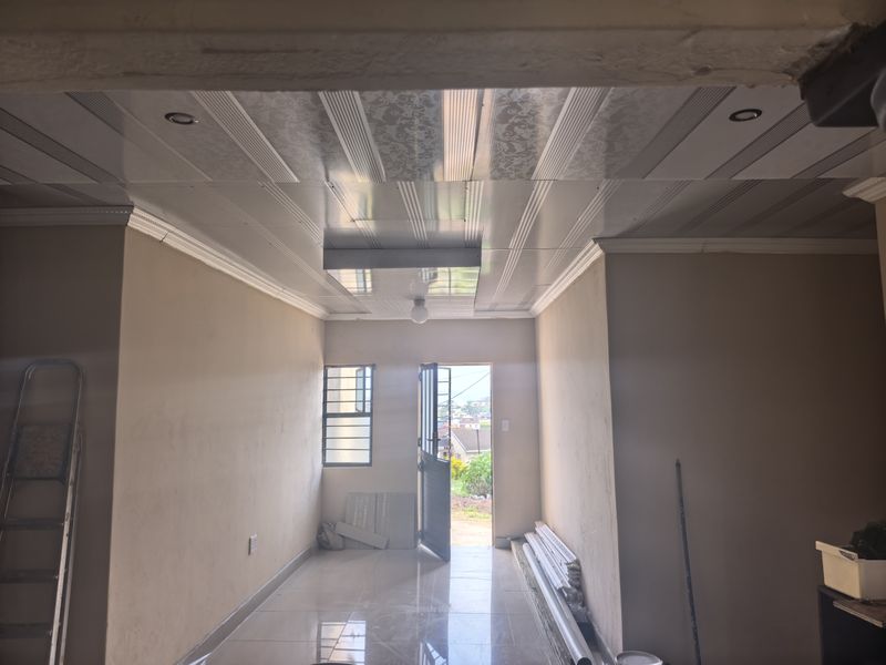 An empty living area featuring decorative PVC ceiling panels, recessed lighting, and tiled flooring, currently undergoing renovation with visible construction materials.