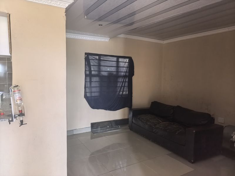 A living area featuring tiled flooring, coved ceilings with plastic paneling, and a window with security bars, showing signs of wear on the walls.