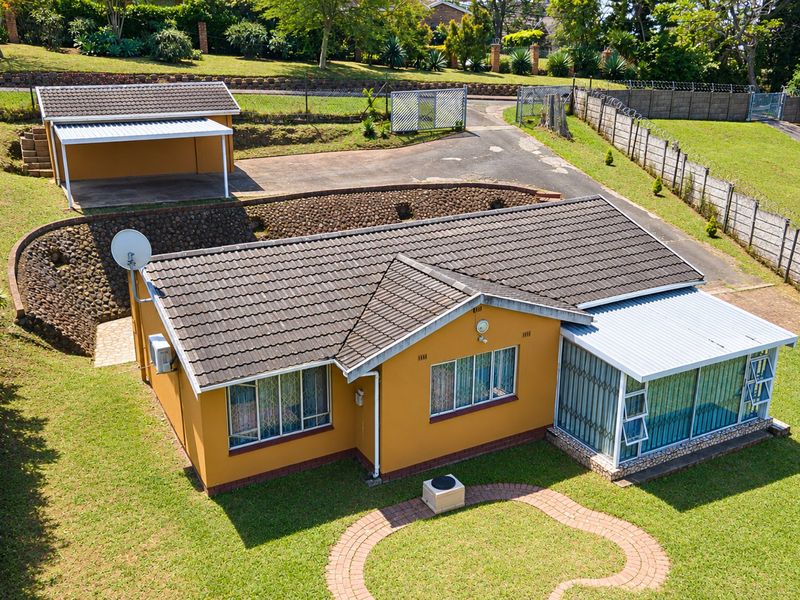 Aerial view of a residential property featuring a tiled roof house, detached carport, paved driveway, and landscaped garden with retaining walls.