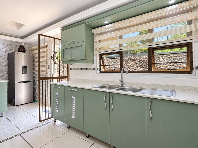 Modern kitchen featuring sage green cabinetry, stone countertops, double basin sink, tiled backsplash, and tiled flooring with a security gate leading outdoors.