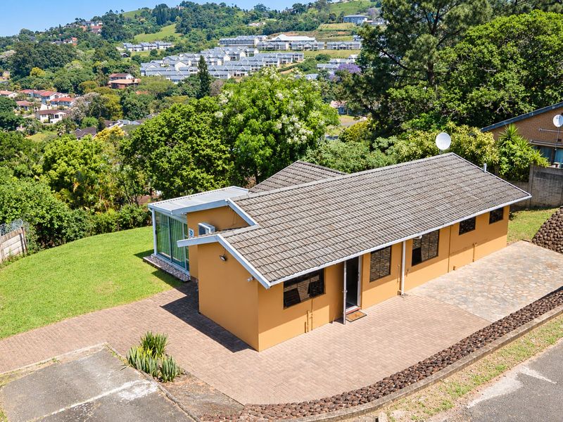 Aerial view of a residential property featuring a tiled roof, paved driveway, and surrounding landscaped garden with scenic hillside views.