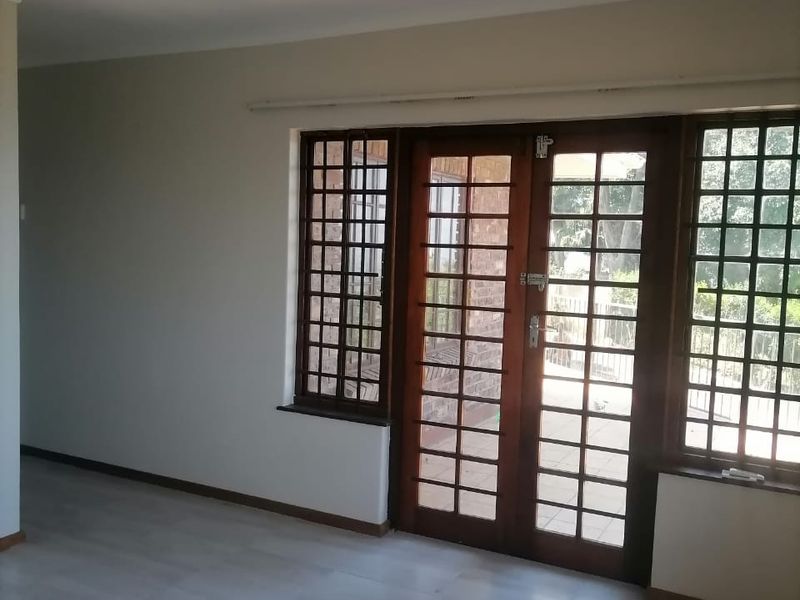 A bright, empty living room featuring tiled flooring, wooden framed glass doors with security bars, and neutral wall finishes.