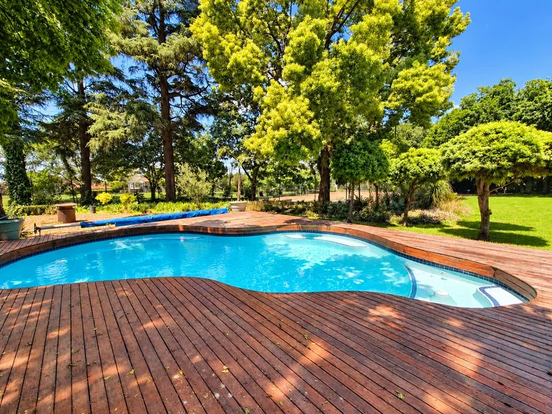 A kidney-shaped swimming pool surrounded by extensive timber decking, set within a lush, mature garden environment.