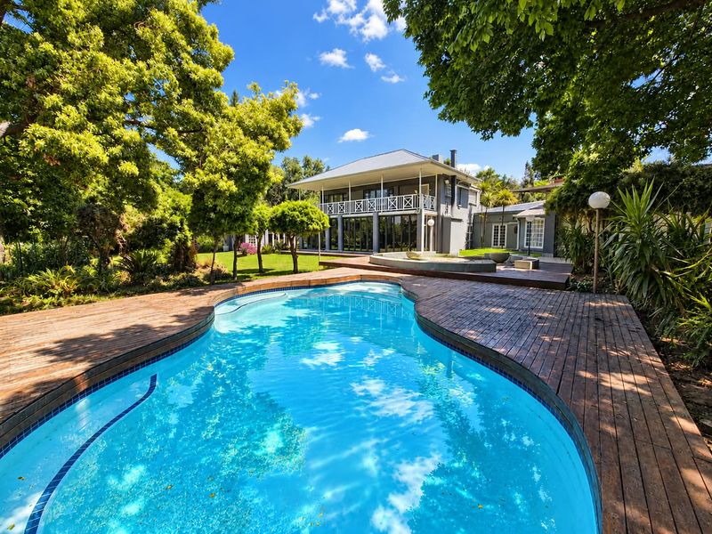 A well-maintained swimming pool area featuring timber decking, lush landscaped gardens, and a view of the two-story residence with a balcony.
