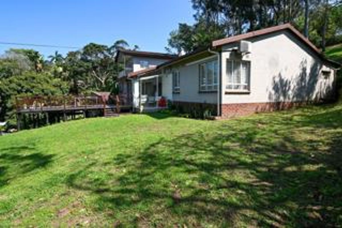 Rear view of the house featuring a raised timber deck, brick base cladding, and a spacious lawn area surrounded by mature trees.