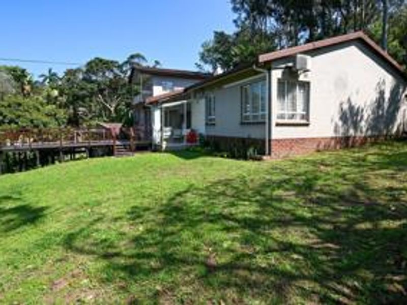 Rear view of the house featuring a raised timber deck, brick base cladding, and a spacious lawn area surrounded by mature trees.