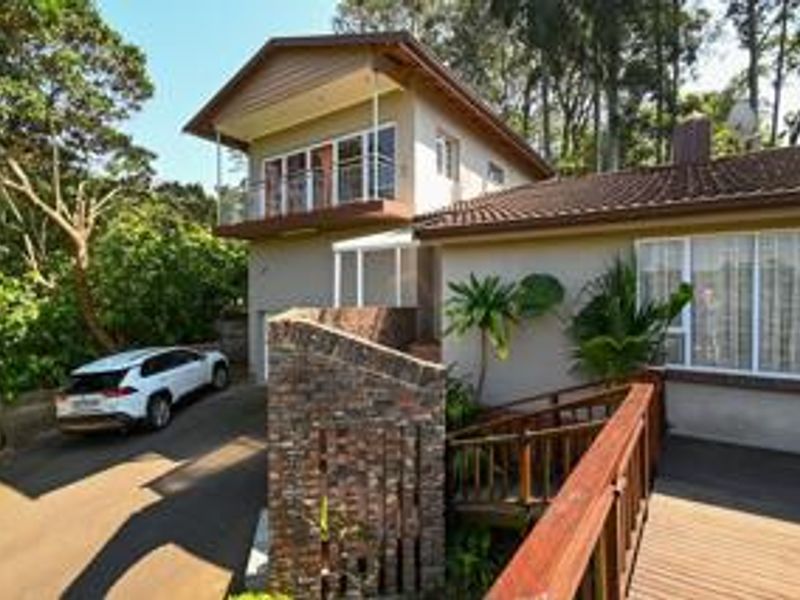 Rear exterior view of a multi-level house featuring a brick retaining wall, timber decking, and an upper-level balcony with sliding glass doors.