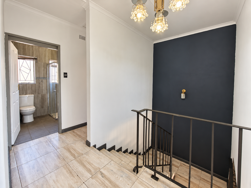 A bright, modern hallway featuring wood-look tiled flooring, a metal staircase balustrade, and a feature dark accent wall, with direct access to a bathroom.