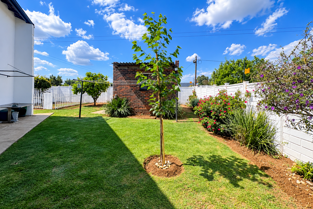 A well-maintained rear garden featuring a manicured lawn, established landscaping, a brick outbuilding, and a paved patio area enclosed by white boundary walls.