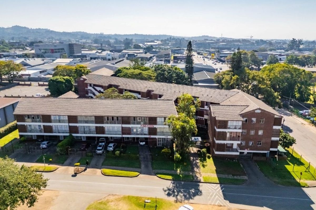 Aerial view of the Pinefern apartment complex, showcasing the multi-story brick building, tiled roofing, and surrounding paved parking areas.