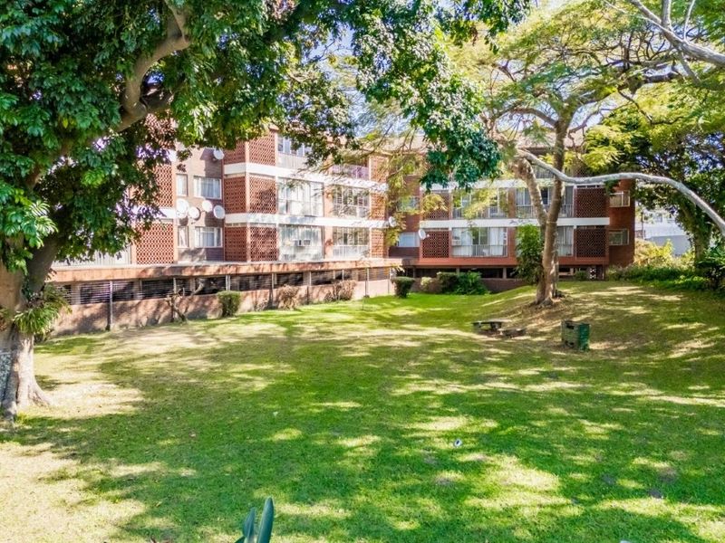 A view of the rear elevation of the apartment building featuring brick facade, private balconies, and a well-maintained communal garden area with mature trees.