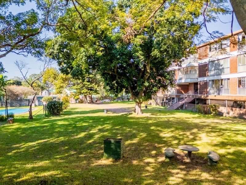 A spacious communal garden area featuring mature trees, lawn, and access to the apartment building and swimming pool.