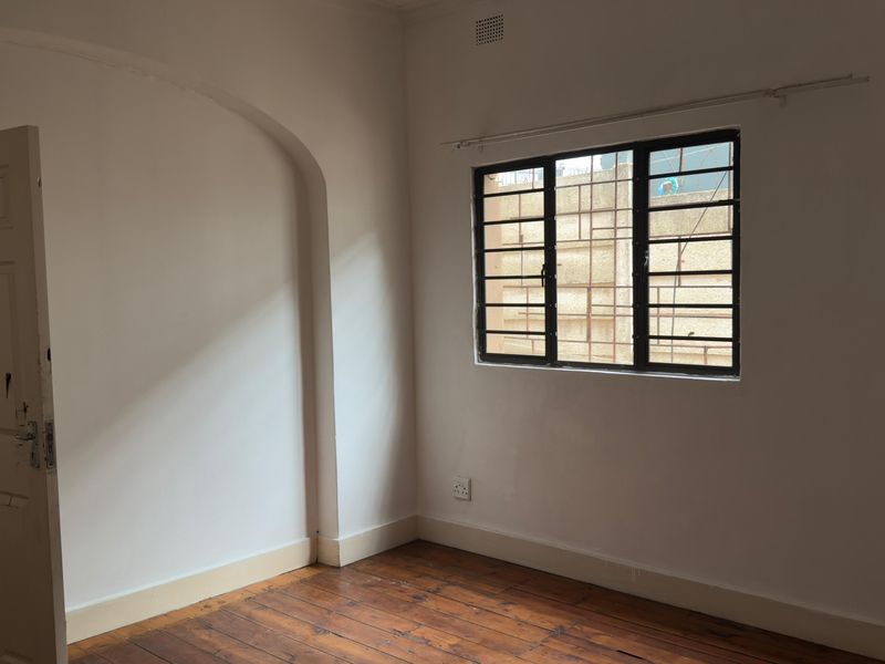 An empty bedroom featuring original timber flooring, an arched architectural detail, and a window fitted with security bars.