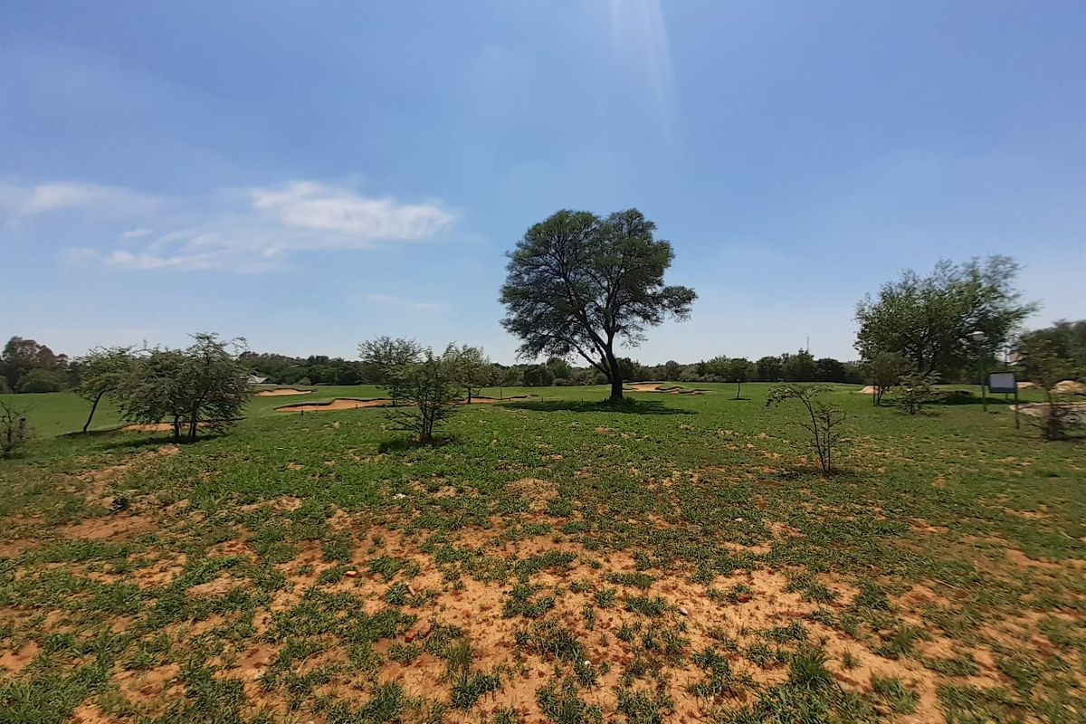 A wide-angle view of a vacant residential plot featuring natural grassland and mature trees under a clear sky.