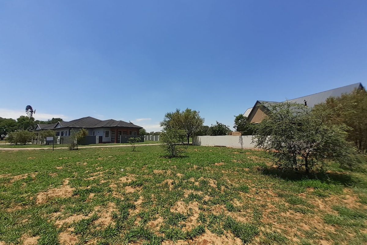 A view of a vacant residential plot featuring natural grass, scattered trees, a boundary wall, and a traditional windmill in the distance.