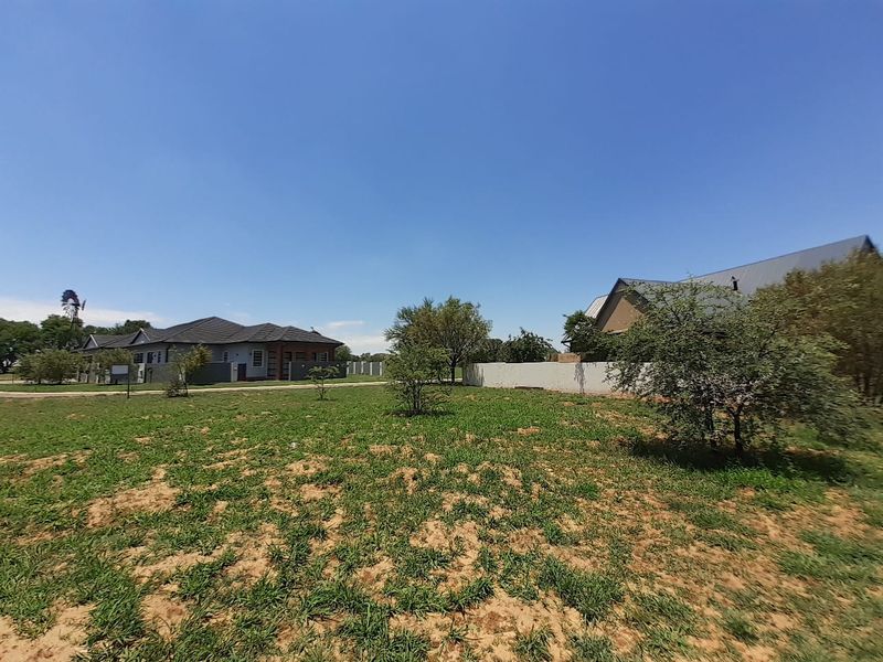 A view of a vacant residential plot featuring natural grass, scattered trees, a boundary wall, and a traditional windmill in the distance.