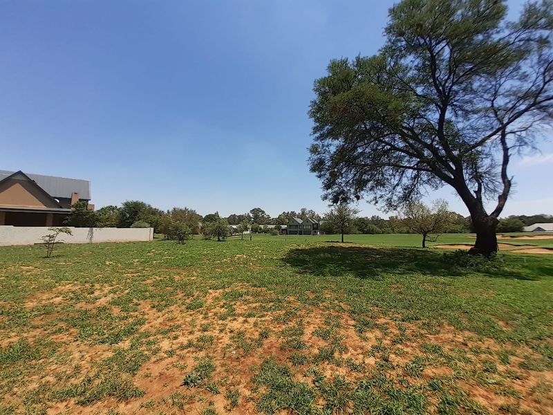 A spacious, open residential plot featuring established trees and a natural grass lawn, offering a clear view of the surrounding estate landscape.