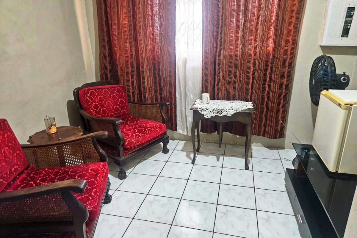 A living room area featuring ceramic floor tiles and a window with a security gate, requiring cosmetic updates to improve the overall finish.