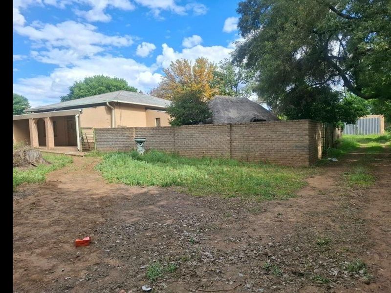 Rear view of the property featuring a brick boundary wall, a thatched roof structure, and a covered patio area with surrounding unpaved garden space.