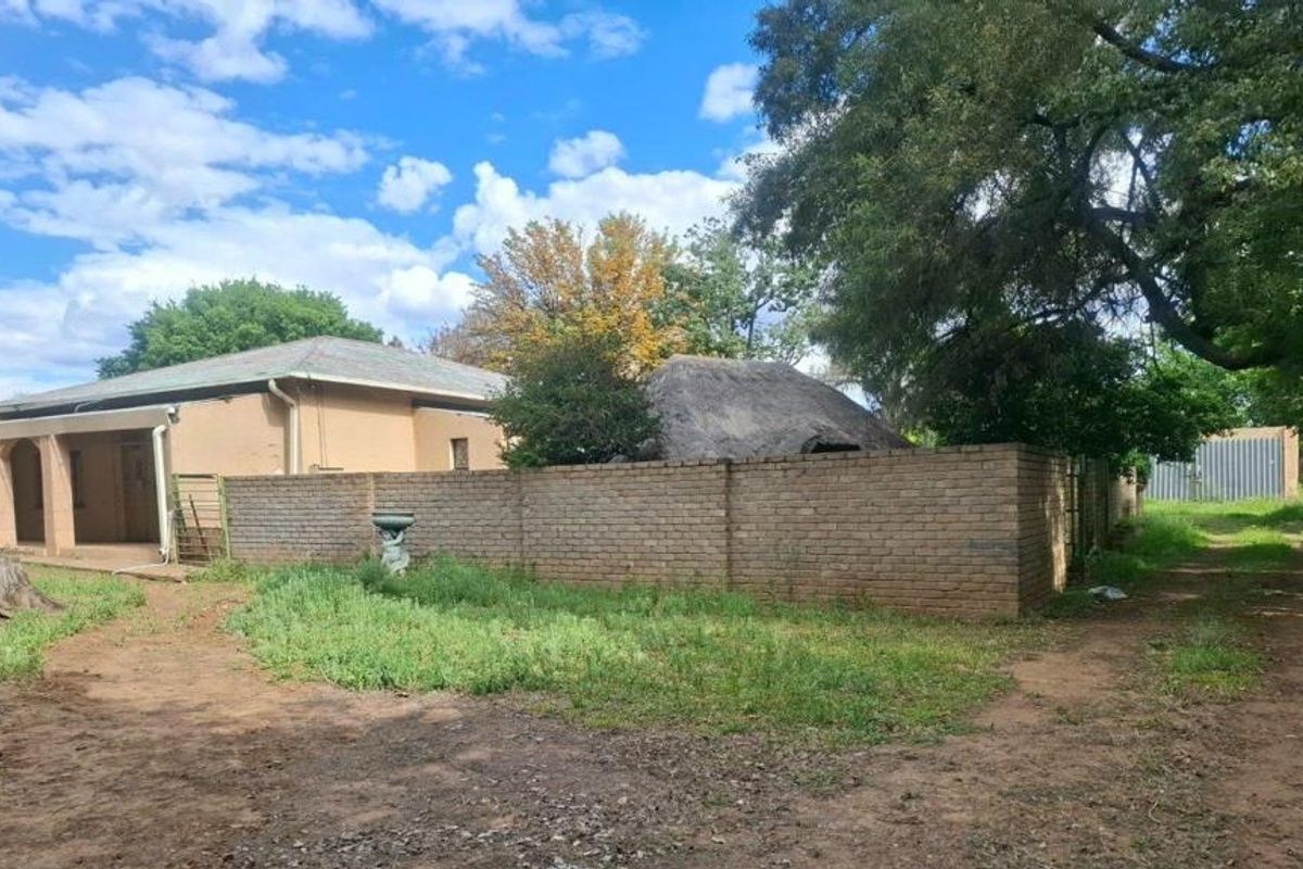 Rear view of the property featuring a brick boundary wall, a thatched roof structure, and a covered patio area with surrounding overgrown garden space.