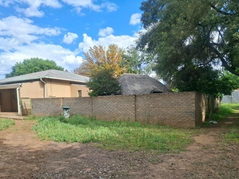 Rear view of the property featuring a brick boundary wall, a thatched roof structure, and a covered patio area with surrounding overgrown garden space.