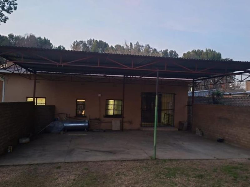 A rear exterior view featuring a large covered patio area with a corrugated metal roof, concrete flooring, and brick boundary walls.