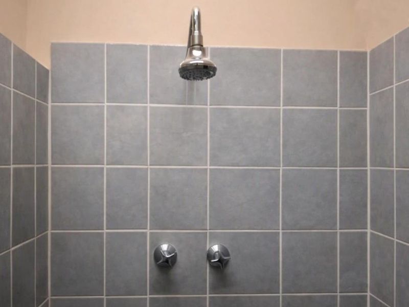 A functional shower enclosure featuring grey wall tiles, a chrome shower head, and dual mixer taps.