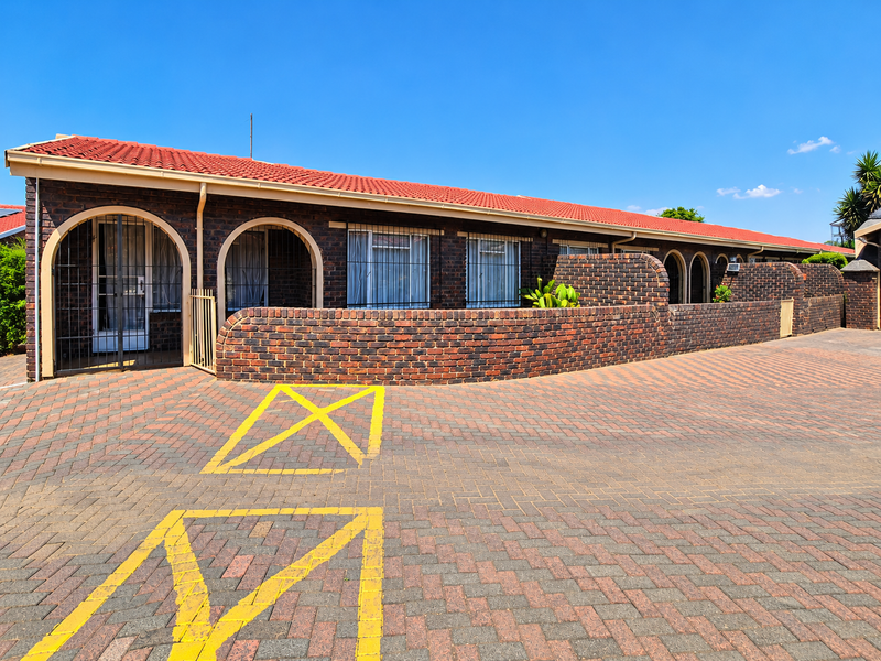 Front exterior view of a townhouse complex featuring a dark brick facade, red tiled roof, arched entryways, and a paved driveway with marked parking bays.