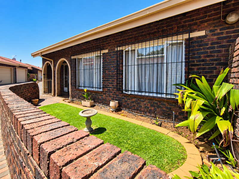 Front view of a face brick townhouse featuring an arched entrance, secure window bars, a small manicured garden, and a low brick boundary wall.