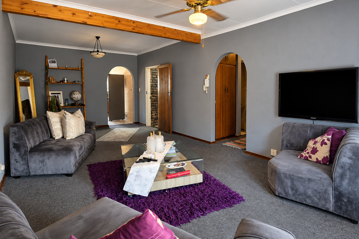A spacious living room featuring grey carpeting, an exposed timber ceiling beam, a ceiling fan, and arched doorways leading to adjacent areas.