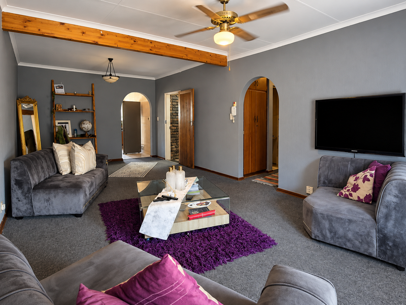A spacious living room featuring grey carpeting, an exposed timber ceiling beam, a ceiling fan, and arched doorways leading to adjacent areas.