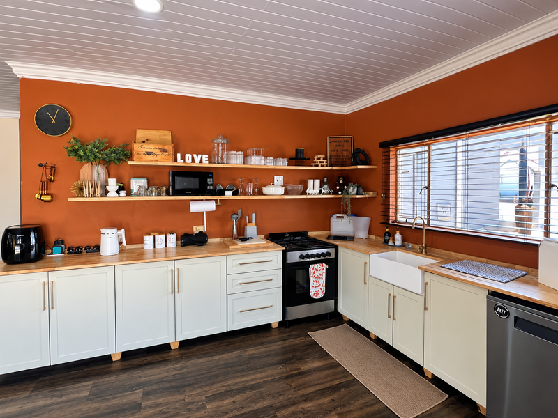 A well-maintained kitchen featuring light-colored shaker cabinetry, butcher block countertops, a farmhouse sink, and open shelving against a vibrant orange accent wall.