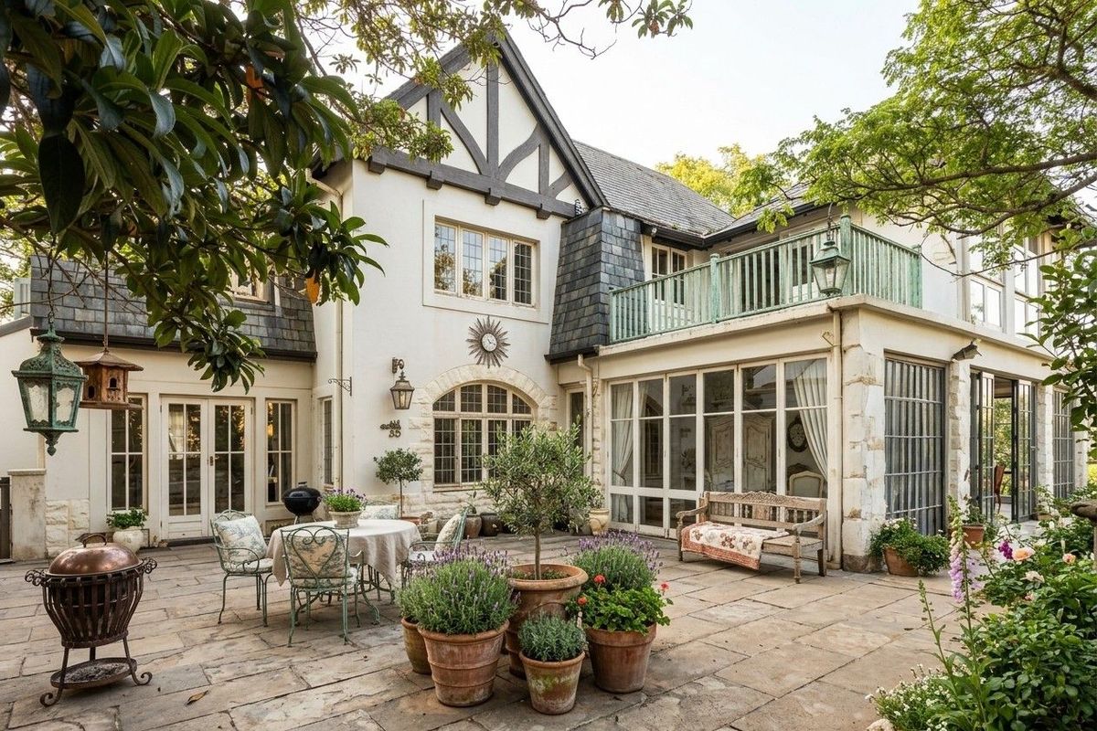 A charming rear exterior view of a multi-story residence featuring a stone-paved patio, timber-framed windows, and an upper-level balcony.