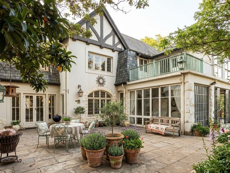 A charming rear exterior view of a multi-story residence featuring a stone-paved patio, timber-framed windows, and an upper-level balcony.