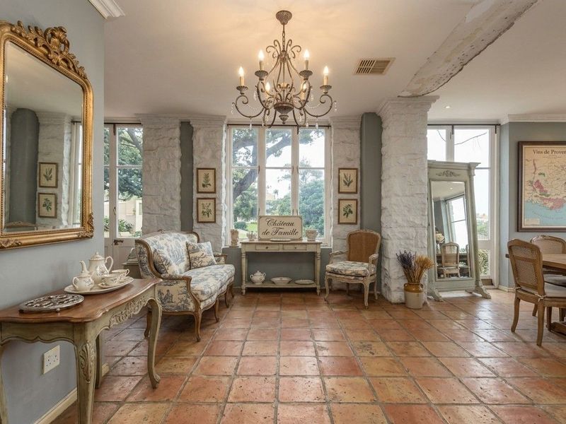 Elegant living area featuring rustic terracotta flooring, exposed stone architectural pillars, and a classic wrought iron chandelier, opening to garden views through french doors.