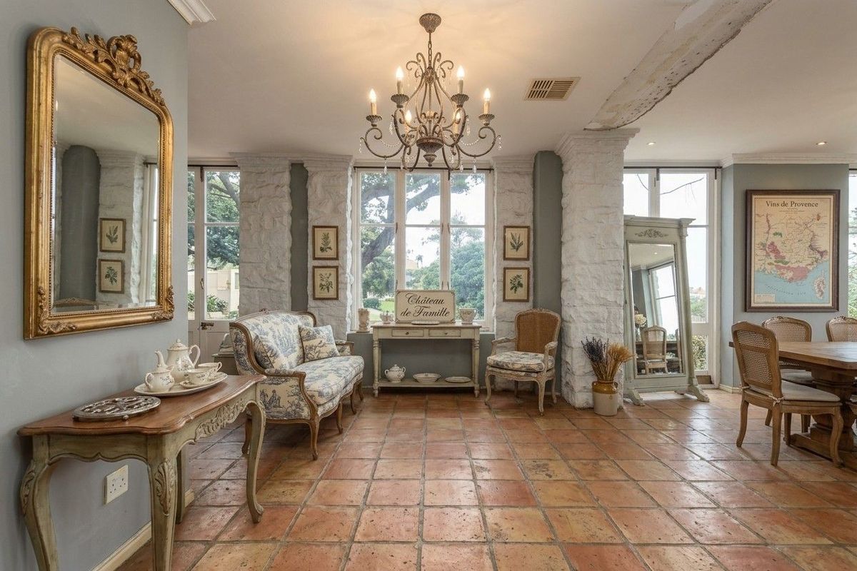 Elegant living area featuring rustic terracotta flooring, exposed stone architectural pillars, and a classic wrought iron chandelier, opening to garden views through french doors.