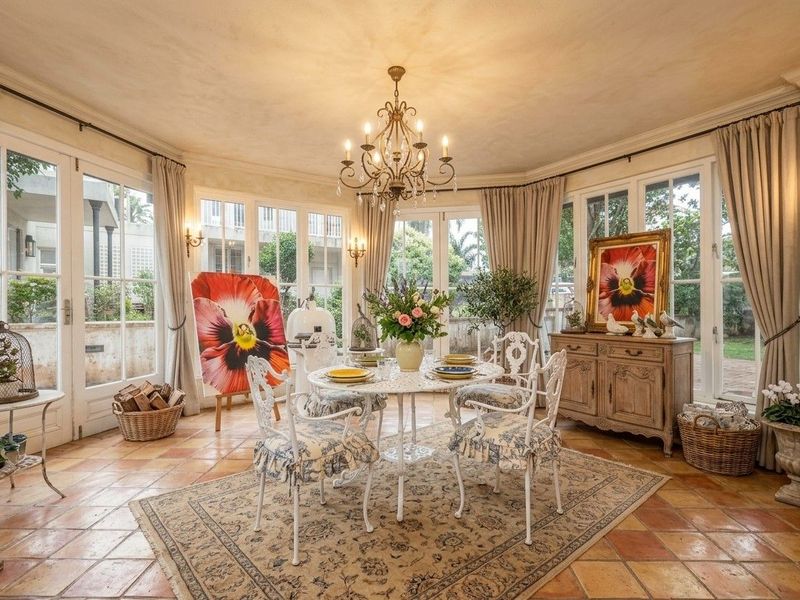 A bright and elegant dining room featuring classic terracotta floor tiles, a decorative chandelier, and multiple sets of French doors providing garden views.