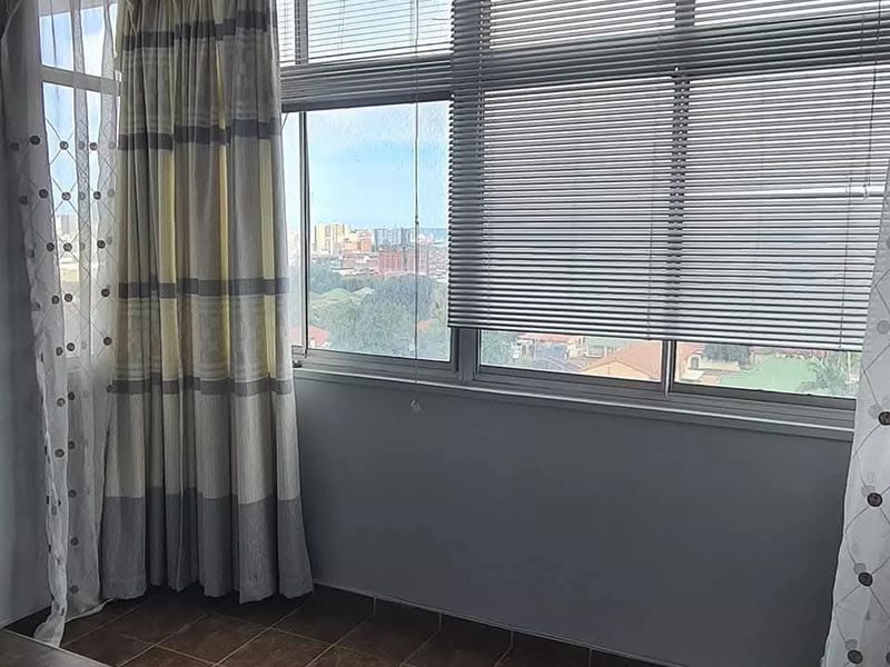 An enclosed balcony or sunroom featuring tiled flooring, large aluminium-framed windows, and venetian blinds, offering an elevated urban view.