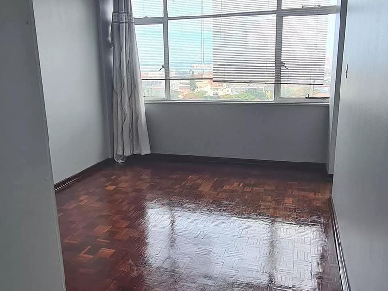A bright, empty bedroom featuring polished parquet flooring and a large window with urban views.