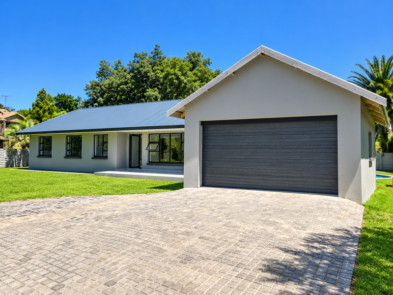 A modern single-story house featuring a metal roof, a paved driveway, and a large garage, set within a well-maintained garden.