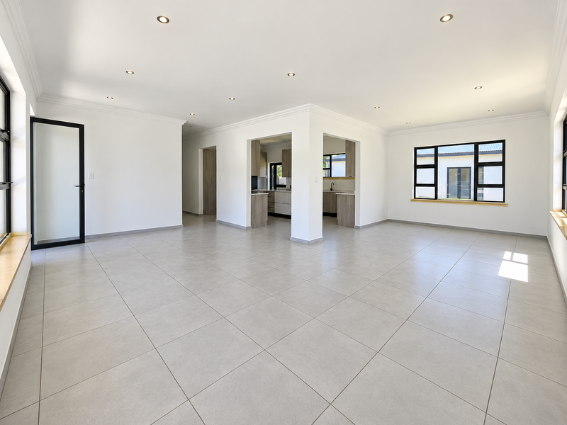 Spacious open-plan living area featuring neutral tiled flooring, recessed downlights, and a modern kitchen with clean lines and black aluminium window frames.