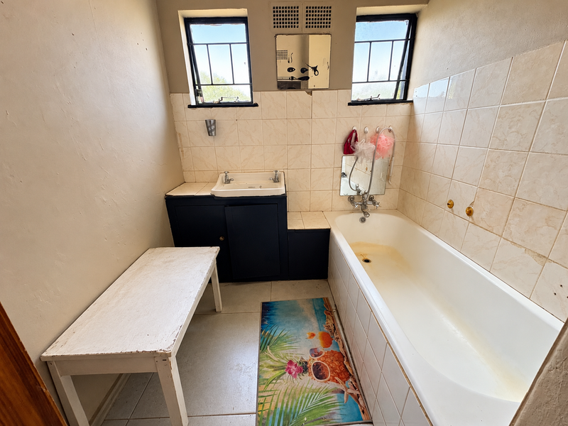 A dated bathroom featuring a built-in vanity with basin, a bathtub with tiled surround, and casement windows, requiring cosmetic updates and fixture refurbishment.