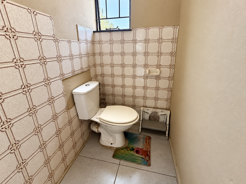 A small, dated bathroom featuring a standard toilet, ceramic wall and floor tiling, and a high-set window with security bars.