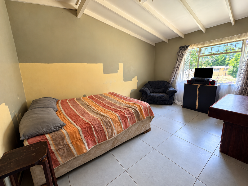 A bedroom featuring tiled flooring, exposed ceiling beams, and a window with security bars. The walls require cosmetic attention and repainting.