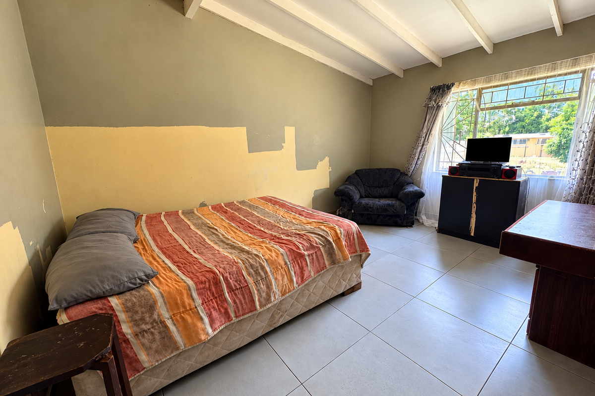 A bedroom featuring tiled flooring, exposed ceiling beams, and a window with security bars. The walls require cosmetic attention and repainting.