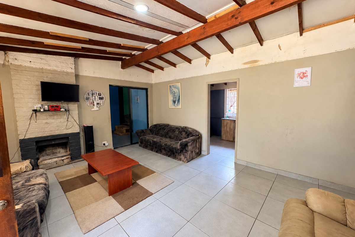 Spacious living room featuring exposed timber ceiling beams, a brick fireplace, and tiled flooring, requiring cosmetic updates to the wall finishes.
