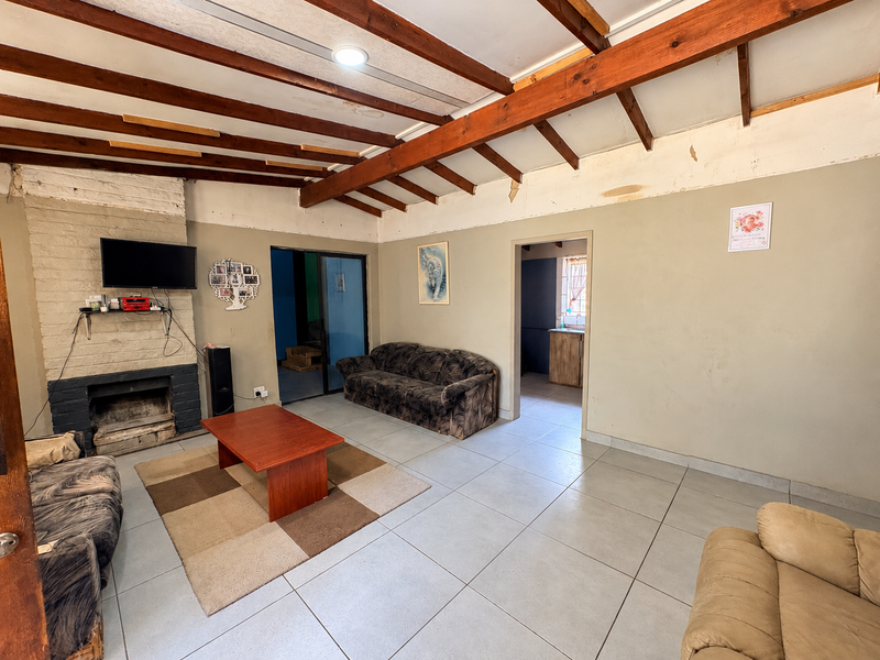 Spacious living room featuring exposed timber ceiling beams, a brick fireplace, and tiled flooring, requiring cosmetic updates to the wall finishes.