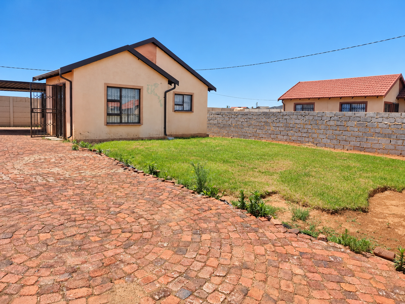 Front view of a single-story house featuring a brick-paved driveway, a lawn area, and a concrete block boundary wall.