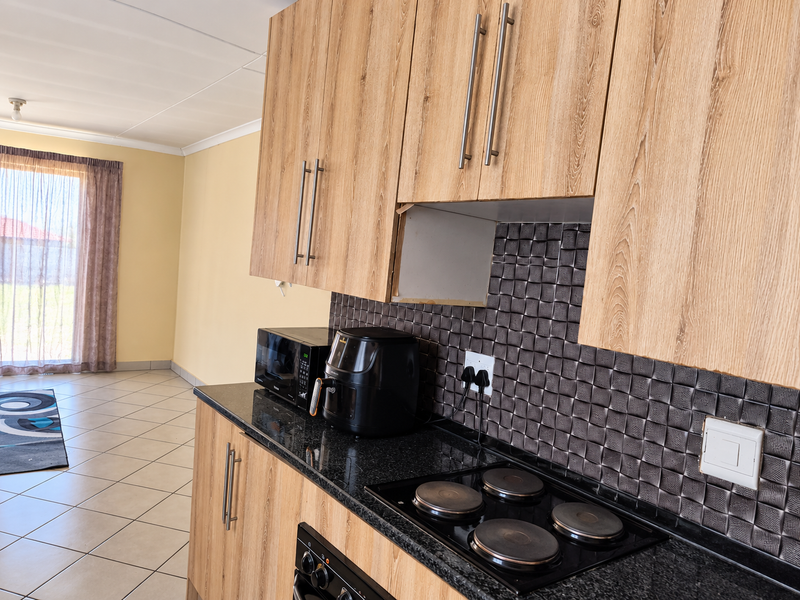 A functional kitchen featuring wood-look cabinetry, black granite countertops, a built-in oven, and a textured mosaic tile backsplash, opening into an adjacent living area.