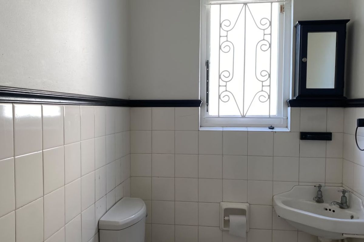 A functional bathroom featuring white wall tiles, a pedestal basin, toilet, and a security-grille window, offering a clean but dated aesthetic.